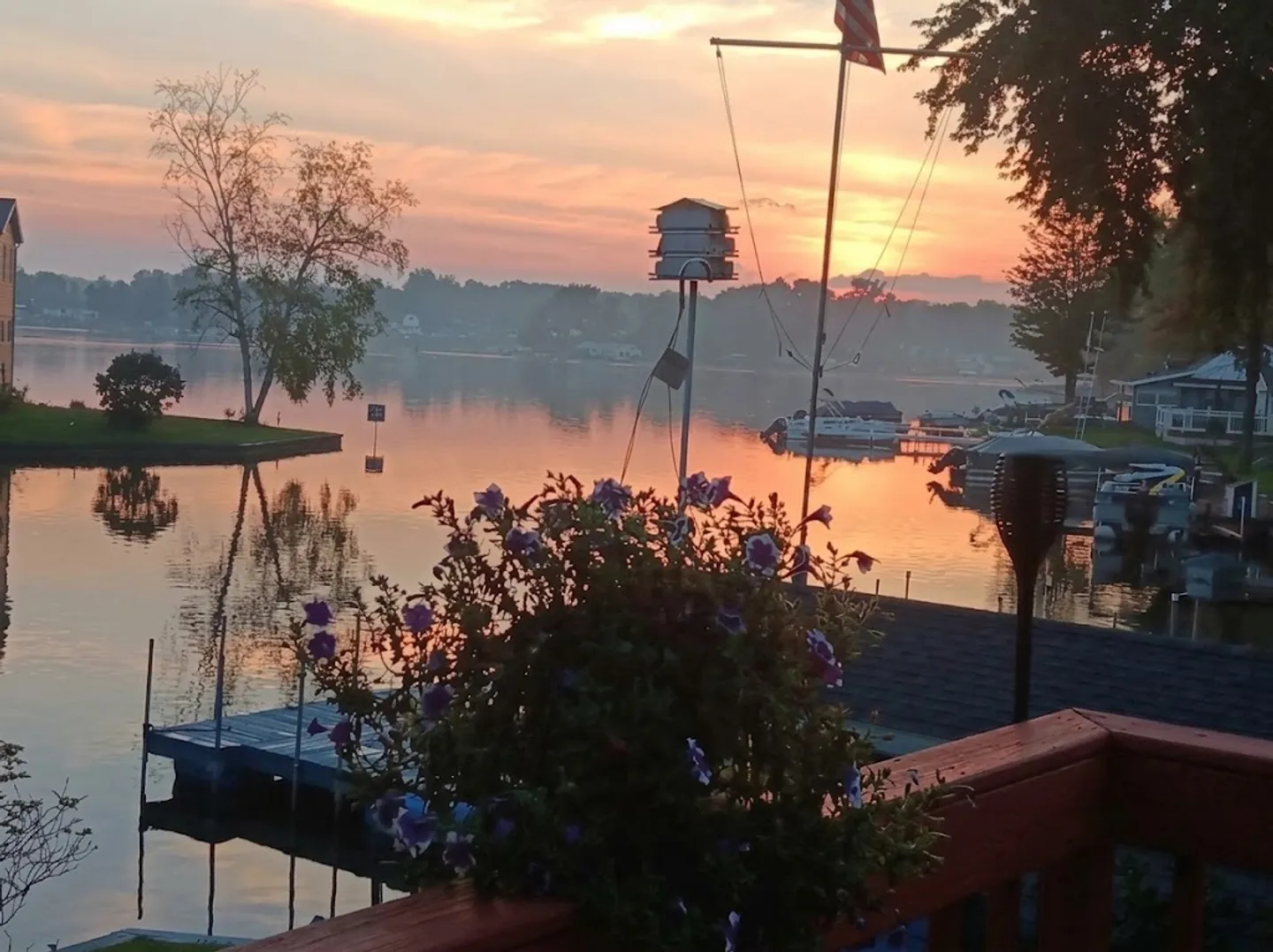 View of a calm lake at sunrise or sunset from a wooden deck railing with a hanging flower basket.