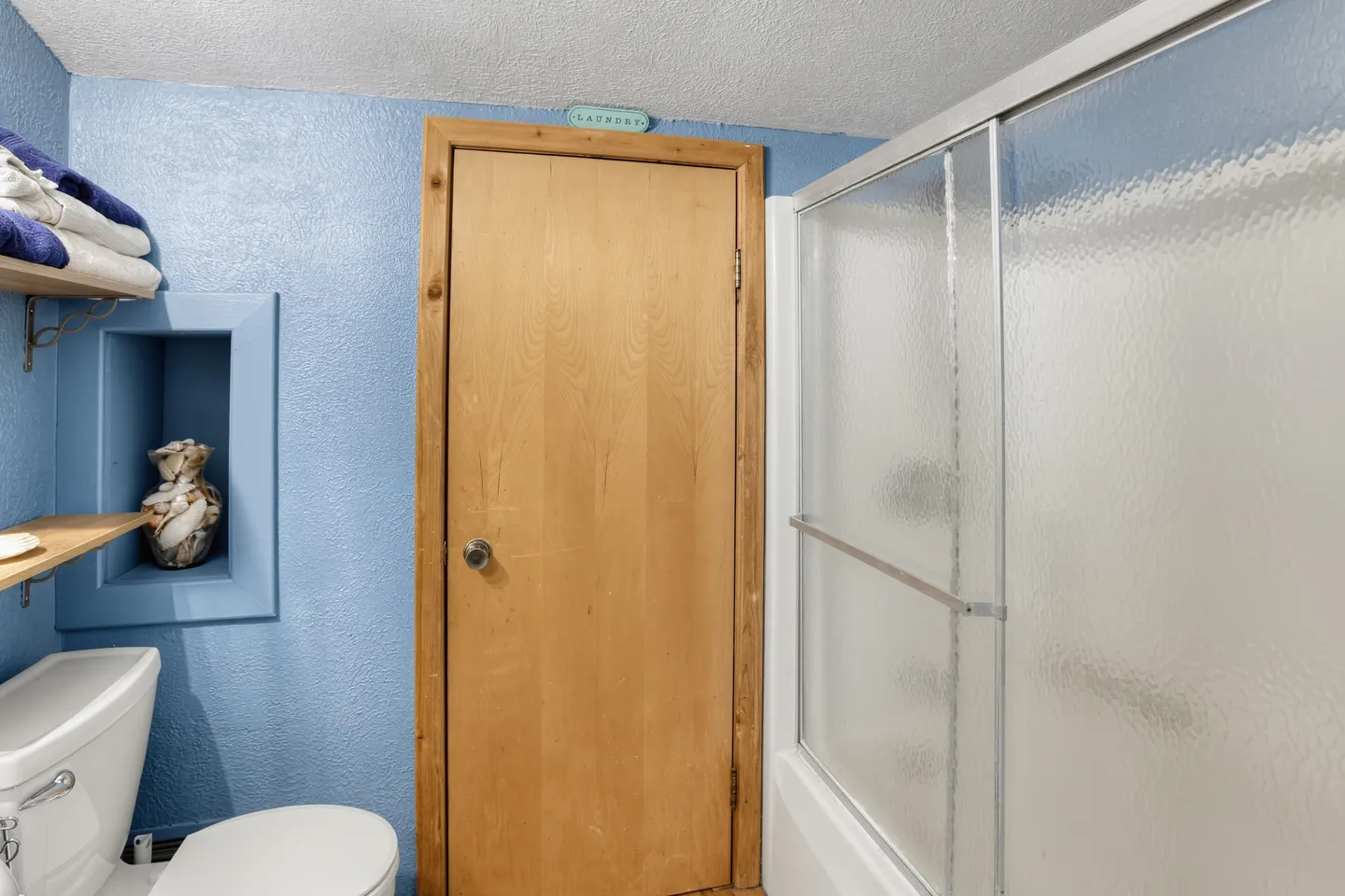 Bathroom showing a white toilet, blue textured walls, a wooden door with a laundry sign, and a shower with frosted sliding glass doors.