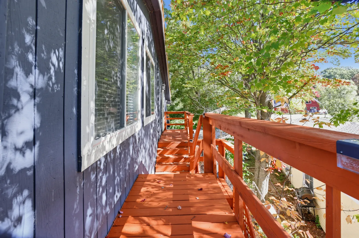A wooden outdoor walkway and stairs with reddish-orange railing along the side of a blue building with trees nearby.