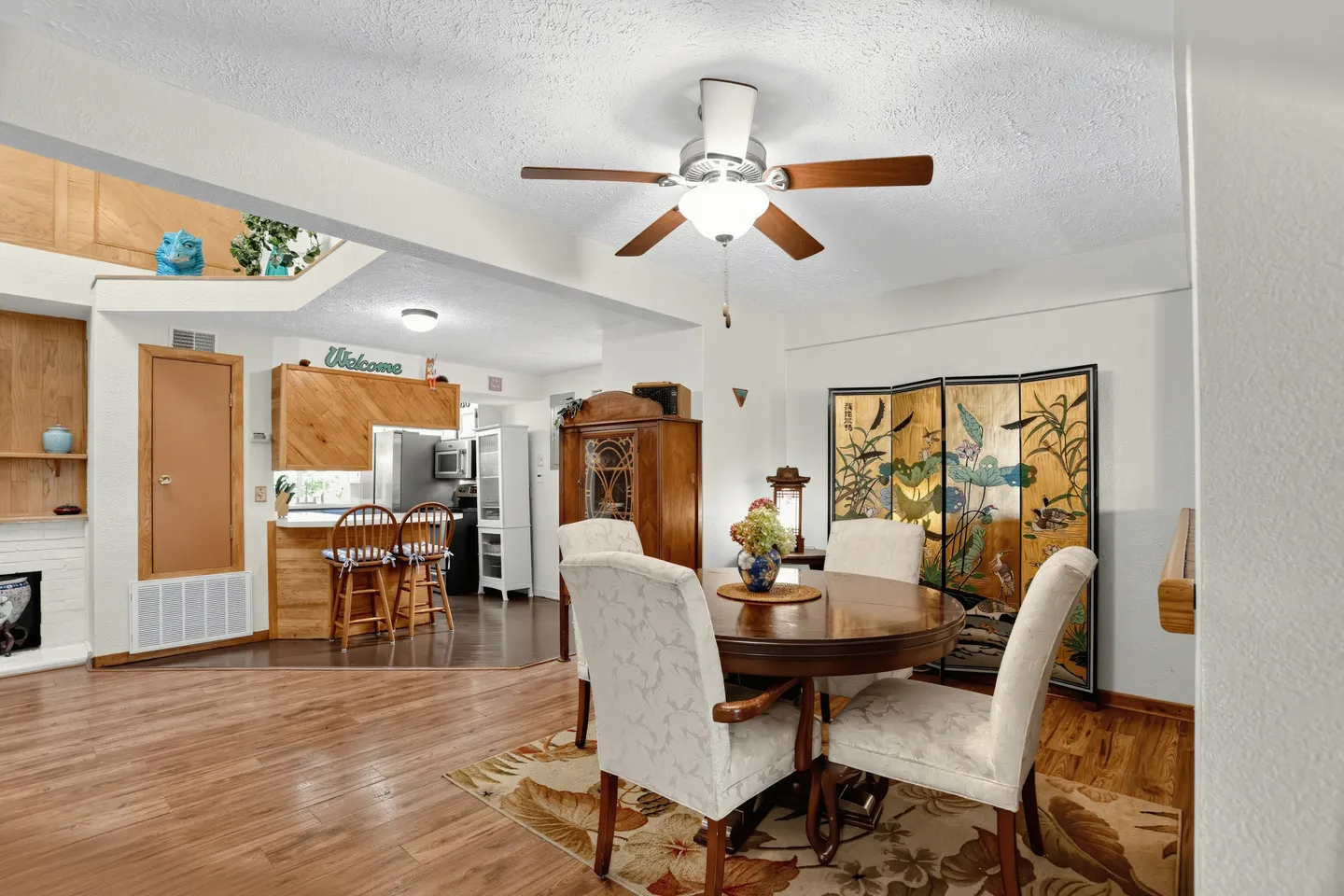 Dining area with a round wooden table, four upholstered chairs, and an overhead ceiling fan looking toward an open kitchen counter.