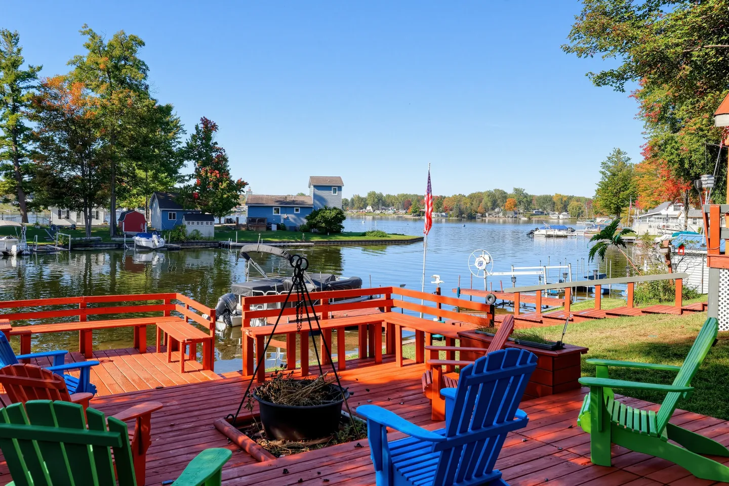 A wooden waterfront deck with red-painted planks, built-in benches, colorful Adirondack chairs, and a direct view of the lake and boat docks.