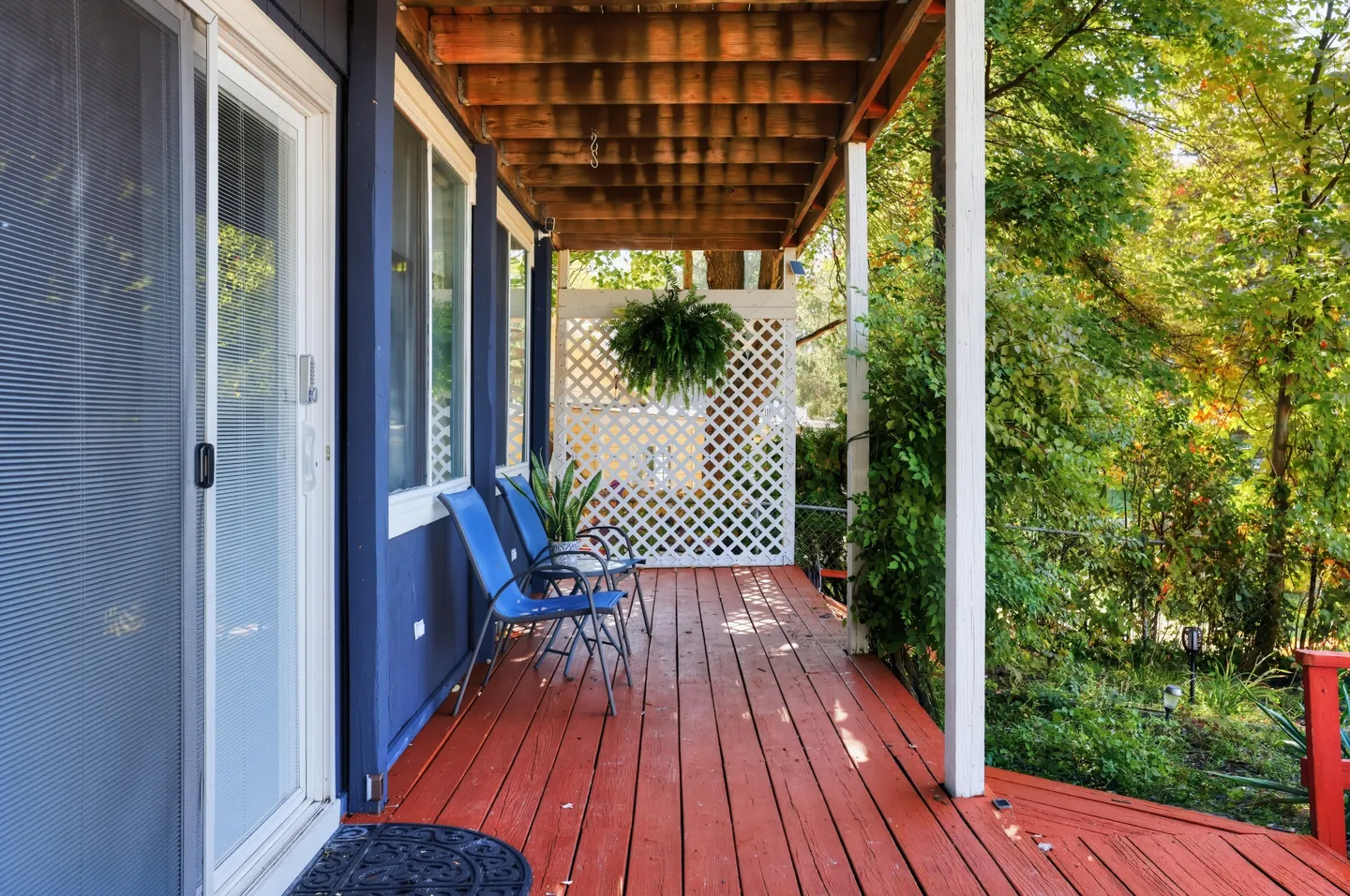A covered outdoor wooden porch with reddish-brown decking, blue siding, two chairs, and a white lattice privacy screen.