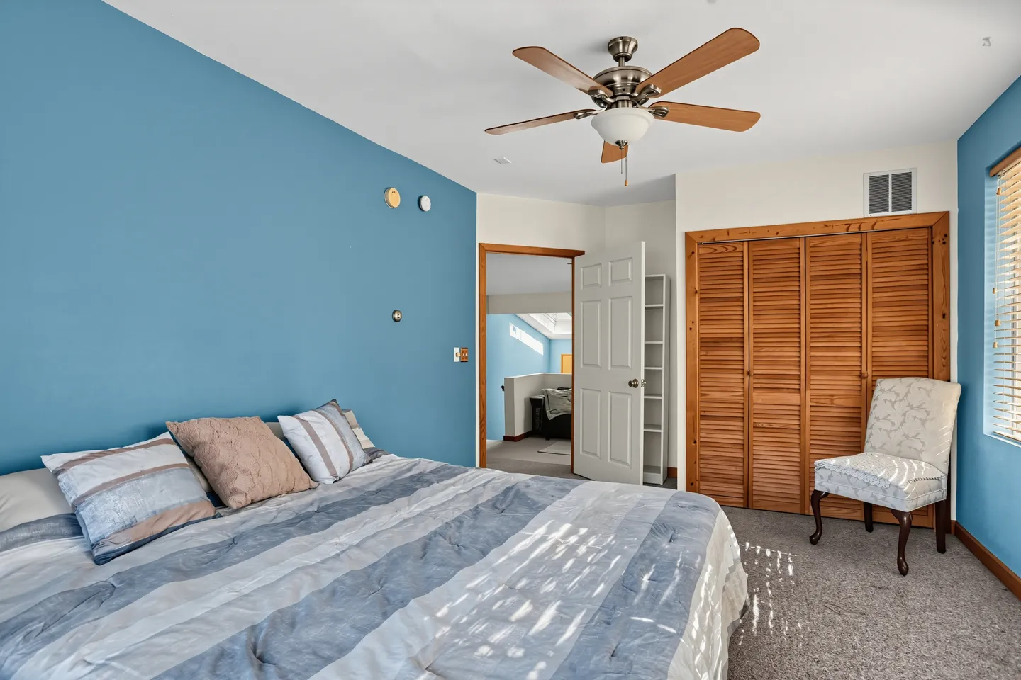 Bedroom featuring a blue accent wall, a bed with a striped comforter, carpet flooring, a ceiling fan, and louvered wood closet doors.