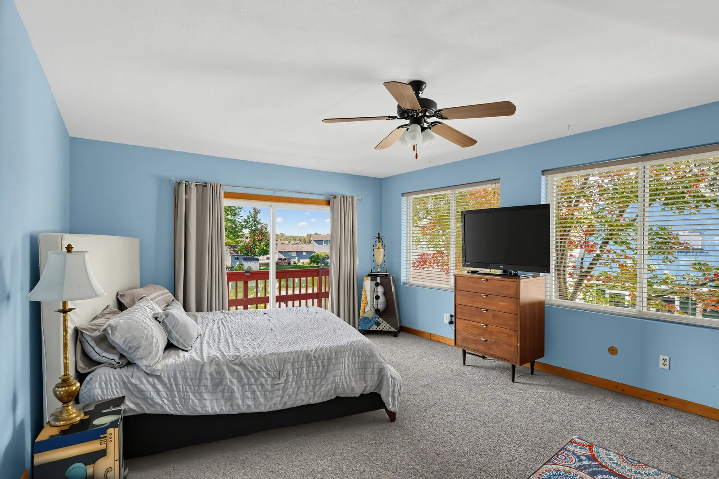 Bedroom with blue walls, carpeted floor, bed with upholstered headboard, ceiling fan, and sliding glass door leading to a balcony.