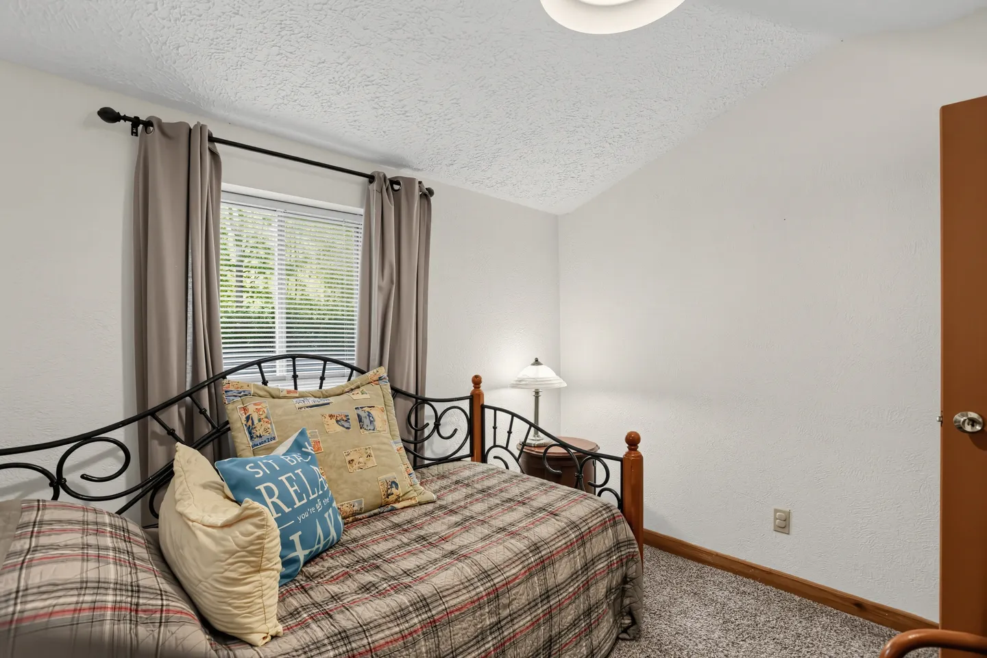 Bedroom with a daybed frame, plaid bedding, carpeted floor, window with curtains, and a nightstand with a lamp.