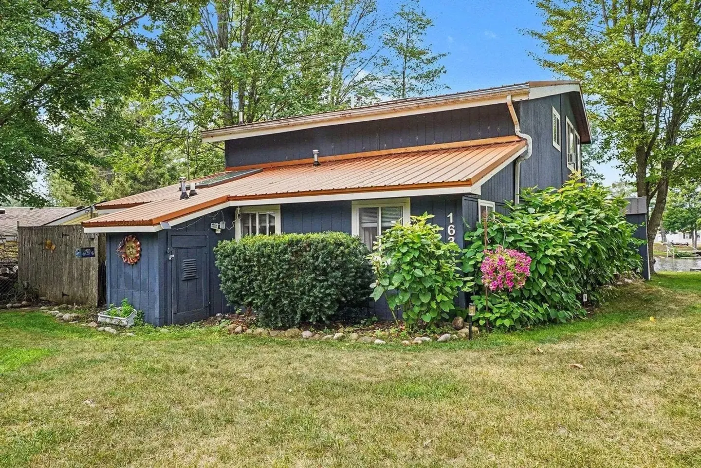 Exterior view of a dark blue house with an orange metal roof, surrounded by green shrubs, trees, and a grassy lawn.