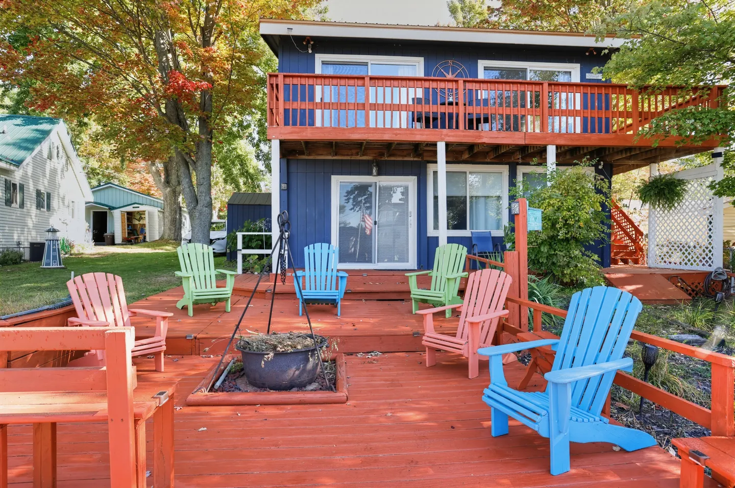 A multi-level reddish-brown wooden deck with colorful Adirondack chairs and a central fire pit in front of a blue two-story house.
