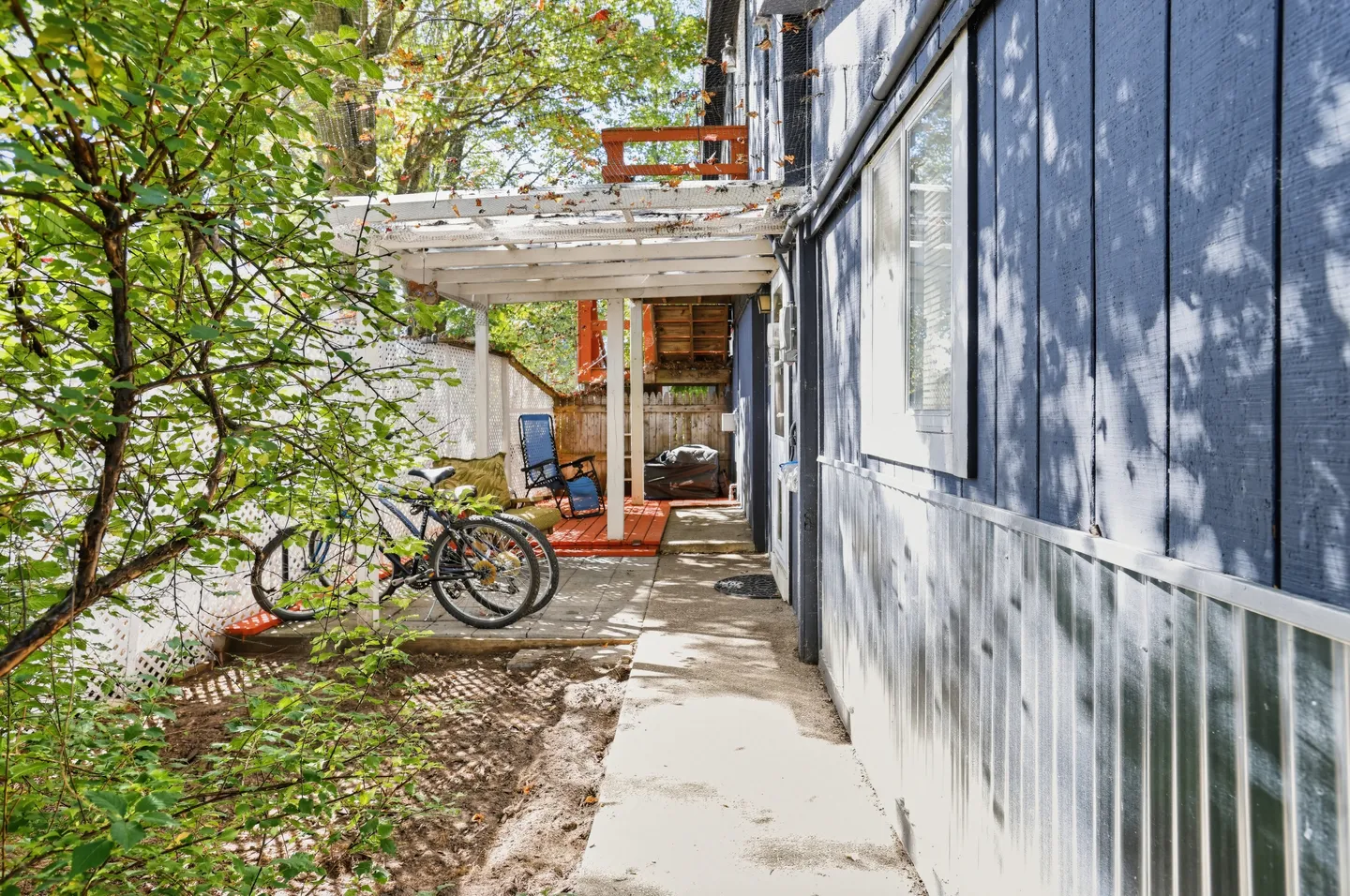 Concrete walkway along the side of a blue building leading to a covered patio area with bicycles and outdoor seating.