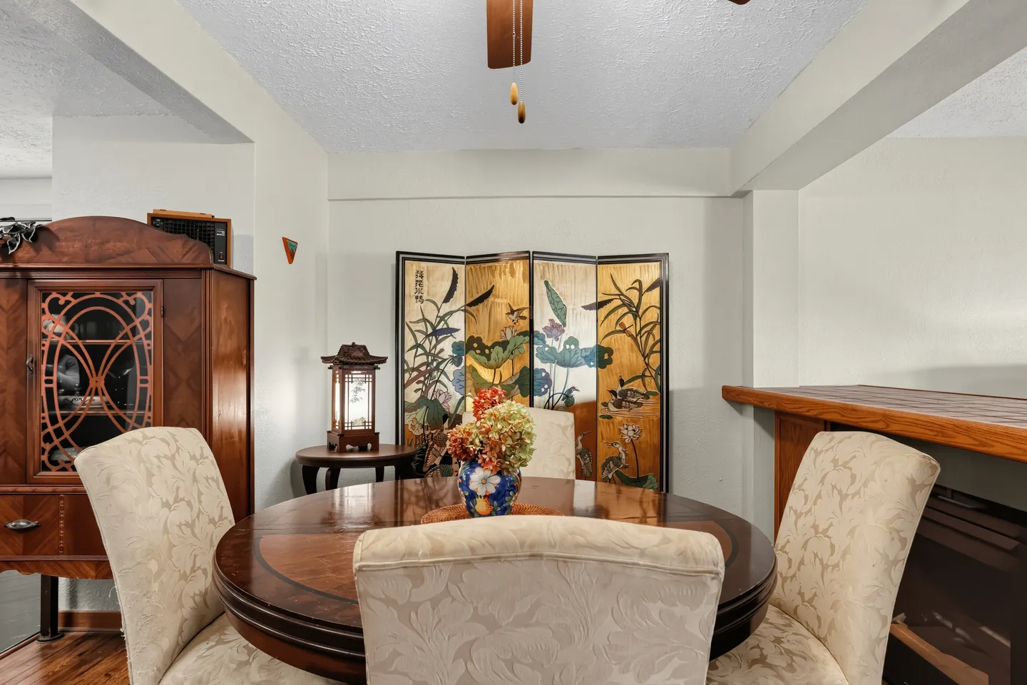 Dining area with a round wooden table, upholstered chairs, an ornate cabinet, and a folding decorative screen against the wall.