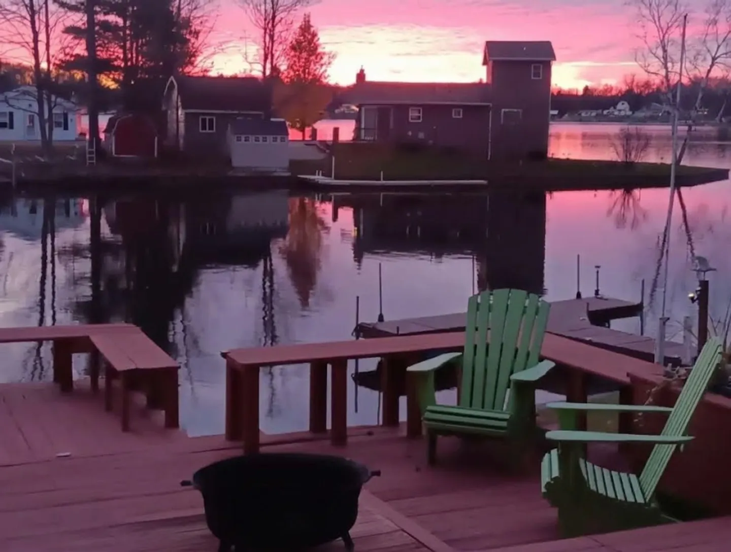 An outdoor wooden deck with green Adirondack chairs, built-in benches, and a fire bowl overlooking calm water and cabins at sunset.