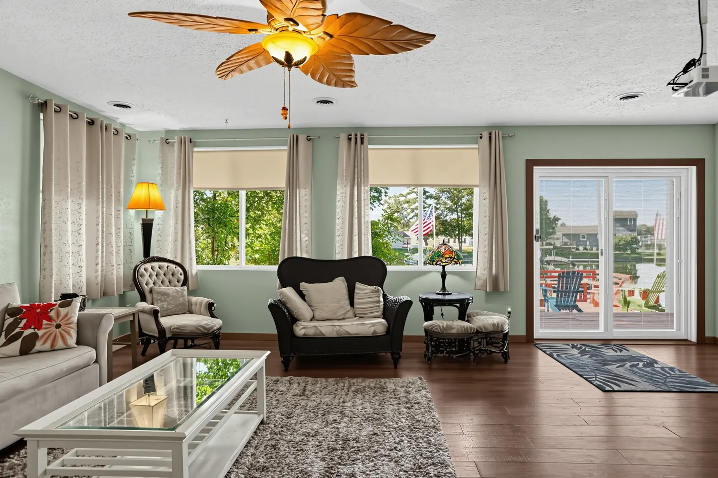 Living room with armchairs, a glass-top coffee table, a palm-leaf style ceiling fan, and sliding glass doors looking out to a waterfront patio.