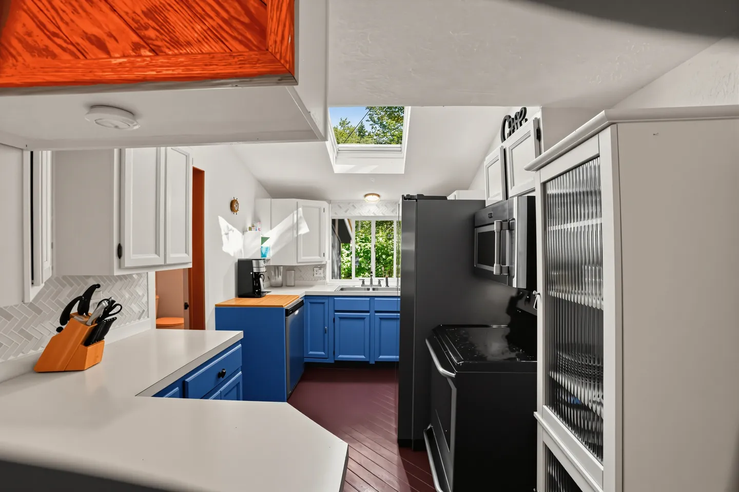A kitchen with bright blue lower cabinets, white upper cabinets, black appliances, and a skylight.