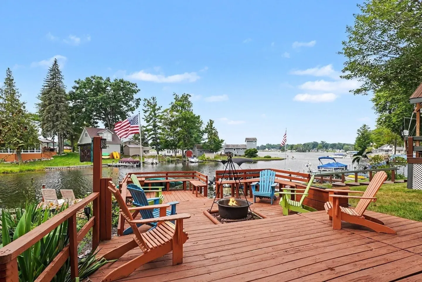 A wooden outdoor deck overlooking a lake with Adirondack chairs and a fire pit.