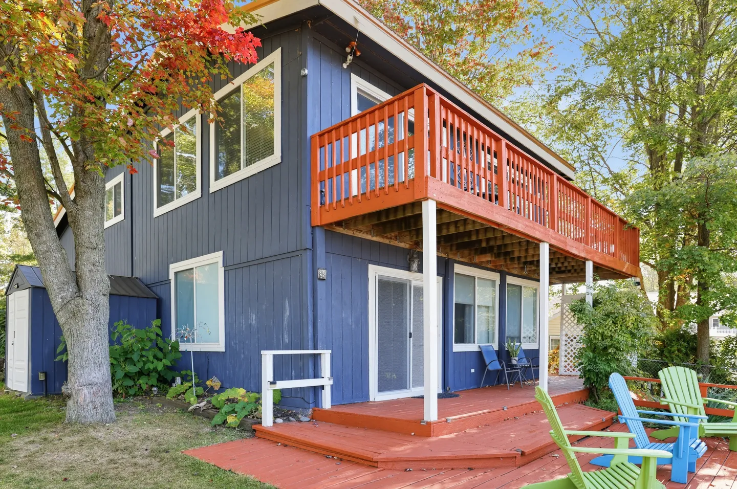Exterior view of a two-story blue house featuring a reddish-brown lower patio deck, an upper balcony with railing, and Adirondack chairs on the lawn.