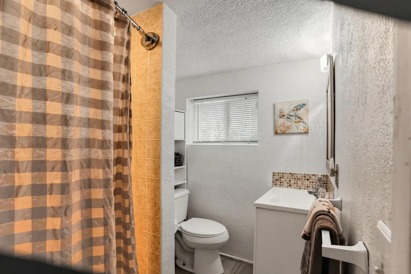 Bathroom showing a toilet beneath a window with blinds, a shower curtain, and a vanity sink with a towel rack.