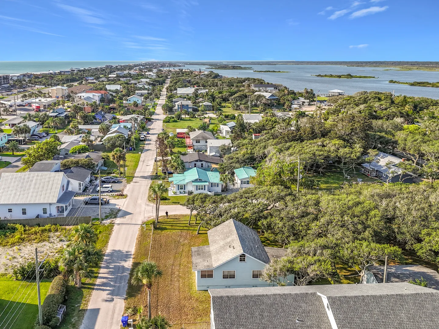 Aerial view of a coastal neighborhood with houses, trees, a straight paved road, and water visible on both sides.