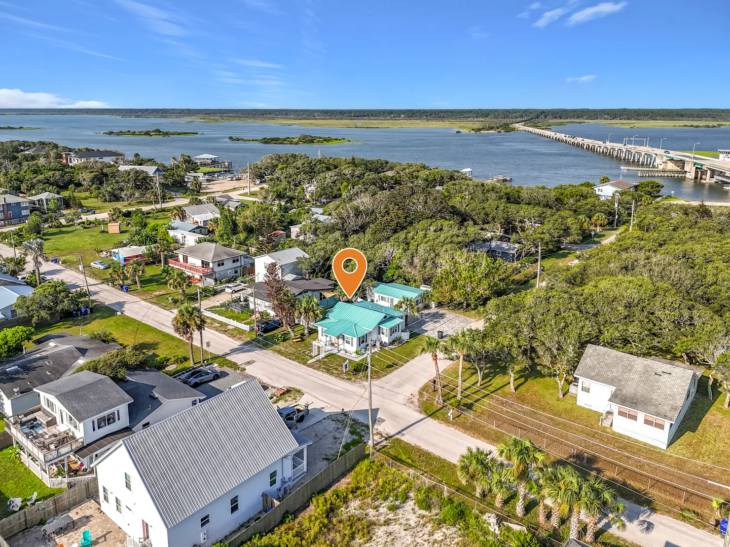 Aerial view of a coastal neighborhood with an orange location marker over a turquoise-roofed house near a river and bridge.