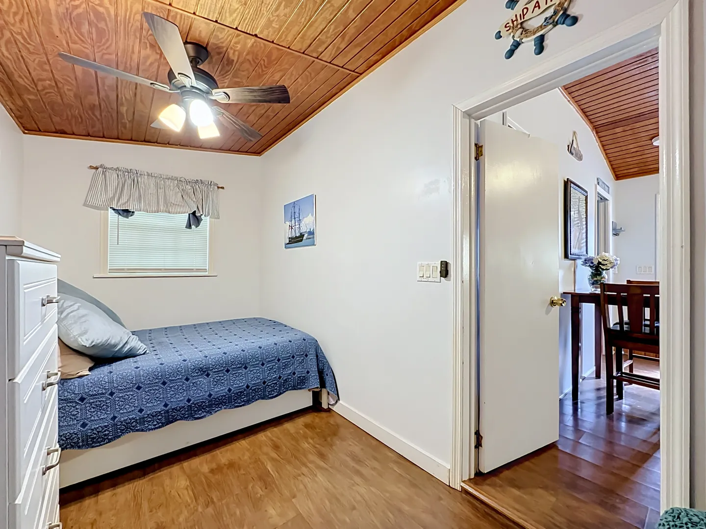 Bedroom with wood-paneled ceiling, ceiling fan, single bed with blue bedspread, and doorway leading to adjacent dining area.