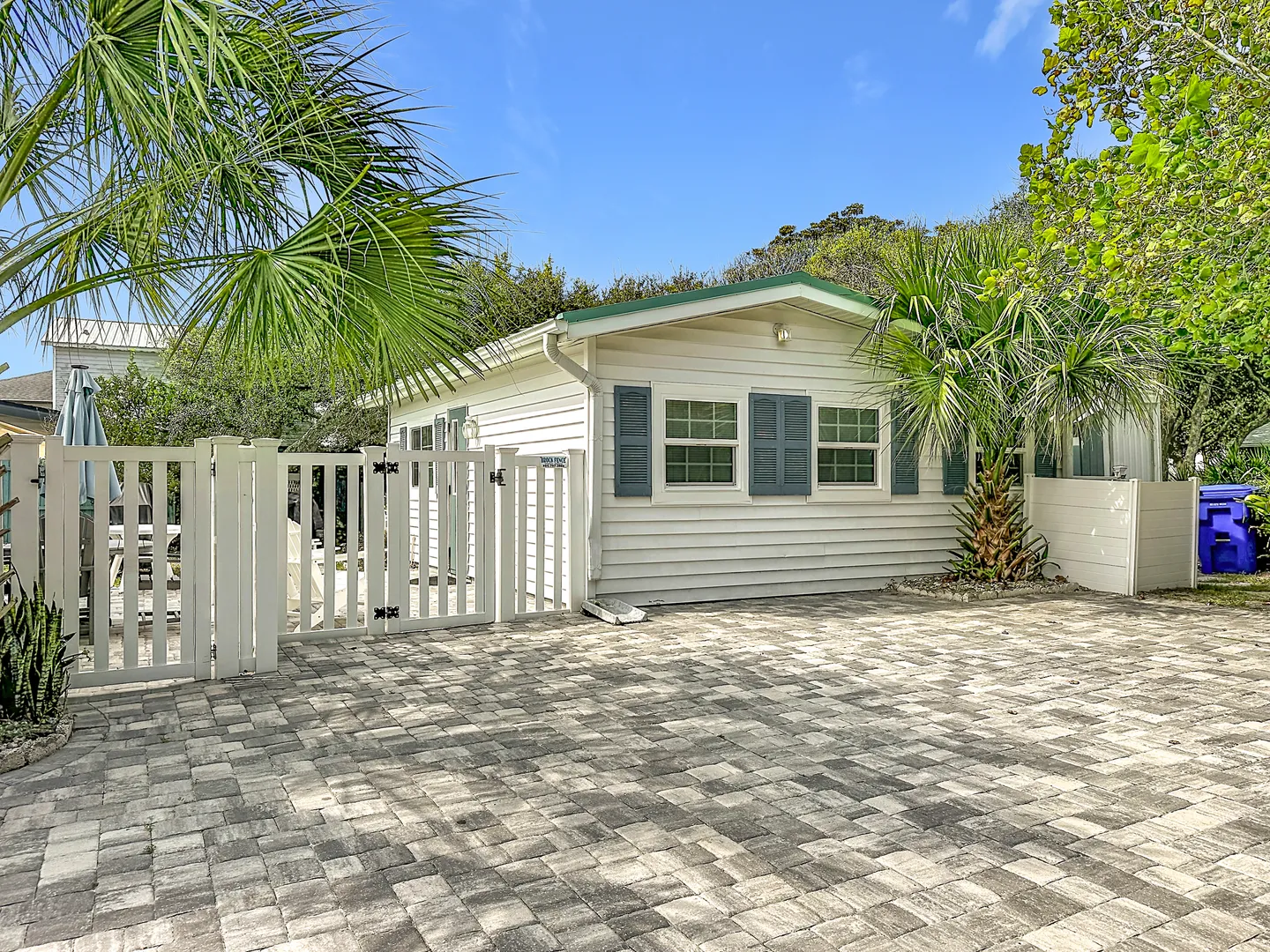 Exterior view of a white siding cottage with blue shutters, a paved driveway, and palm trees under a blue sky.