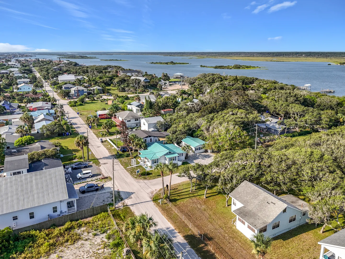 Aerial view of a coastal neighborhood with single-family houses, paved roads, lush trees, and a wide body of water in the background.