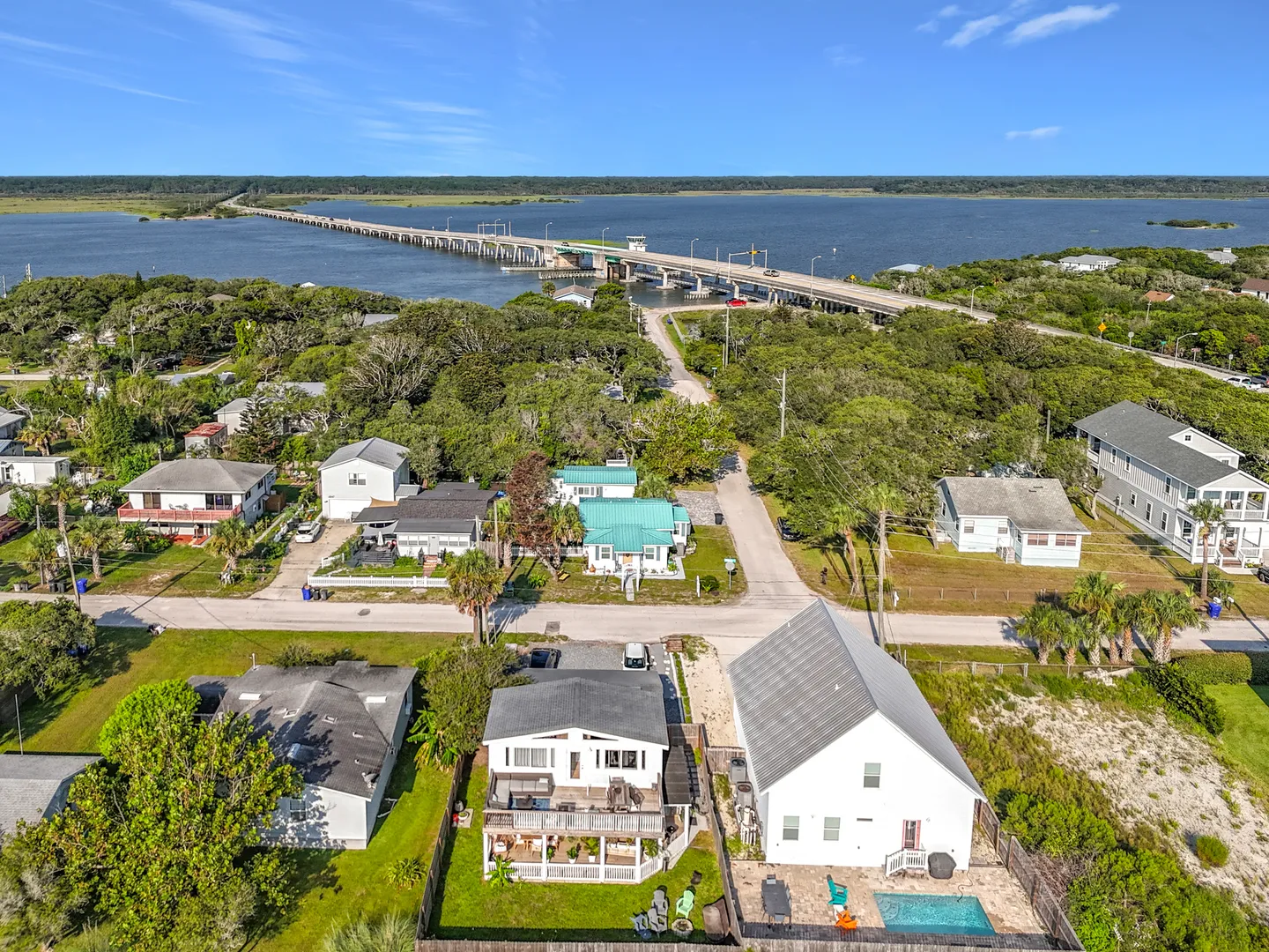 Aerial view of a coastal neighborhood featuring houses, yards with decks and a pool, near a long bridge crossing a wide body of water.