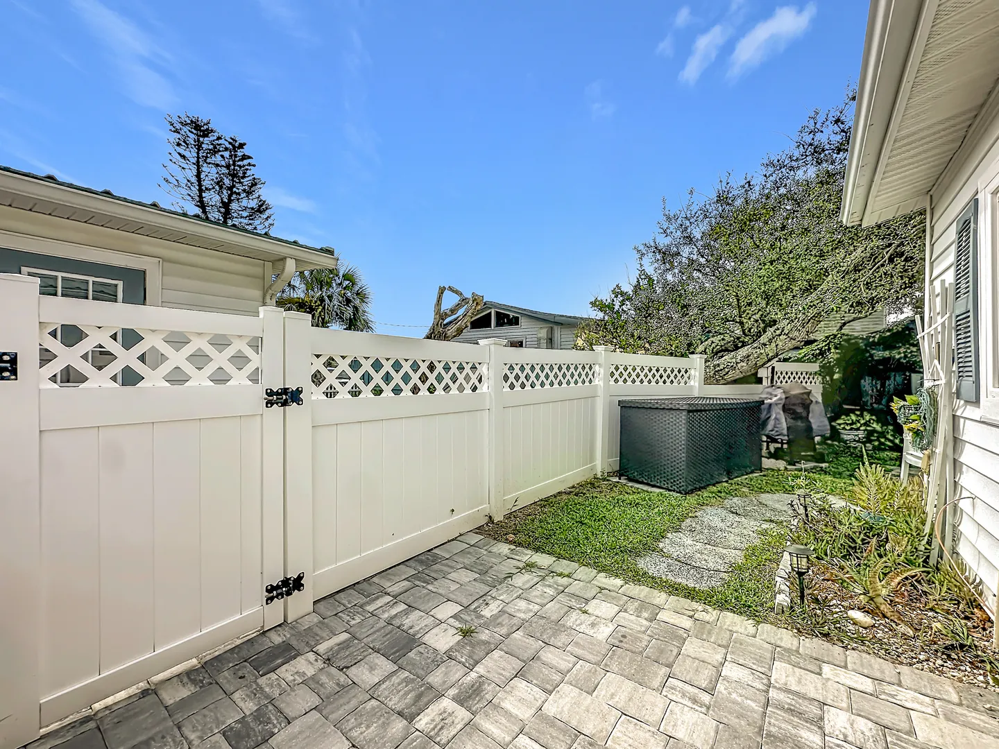 A fenced side yard with stone pavers, a small grass strip with stepping stones, a storage box, and a white lattice-topped fence under a blue sky.