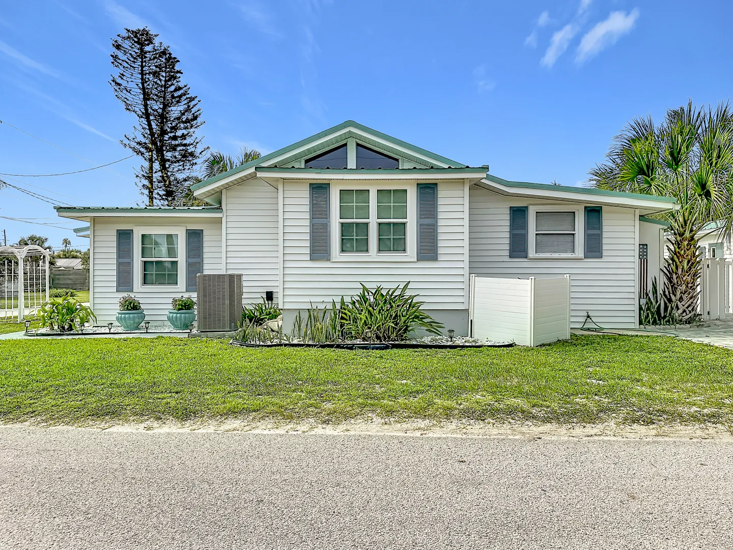 Front exterior of a single-story white home with blue shutters, a green roofline trim, and a small front lawn under a blue sky.
