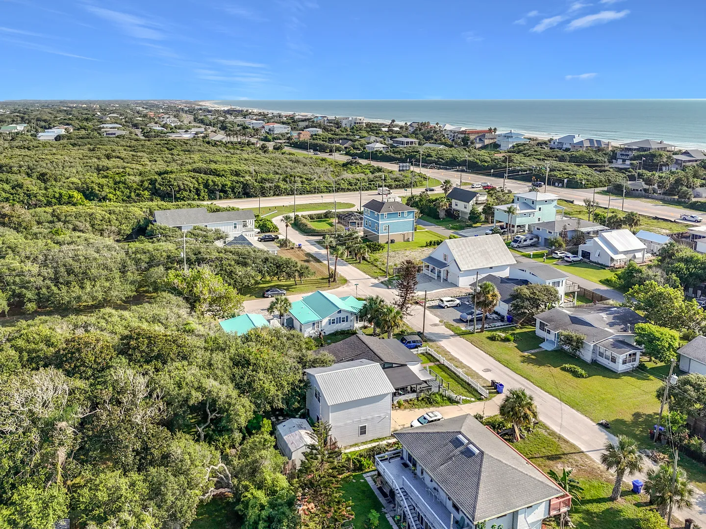 Aerial view of a coastal neighborhood with residential homes, lush greenery, and the ocean in the background under a clear sky.