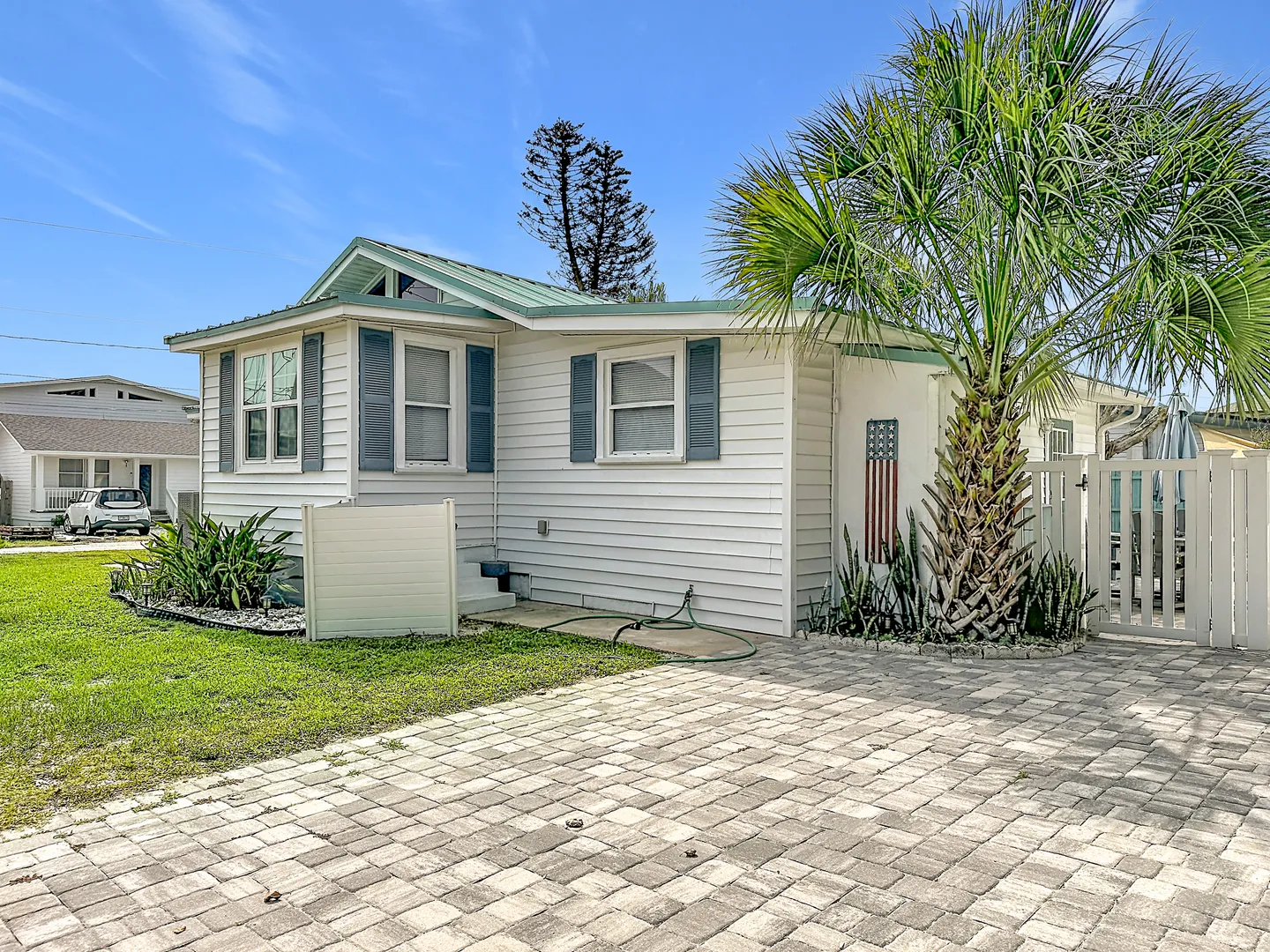 Exterior view of a single-story white house with blue shutters, a paved driveway, and a palm tree under a bright blue sky.