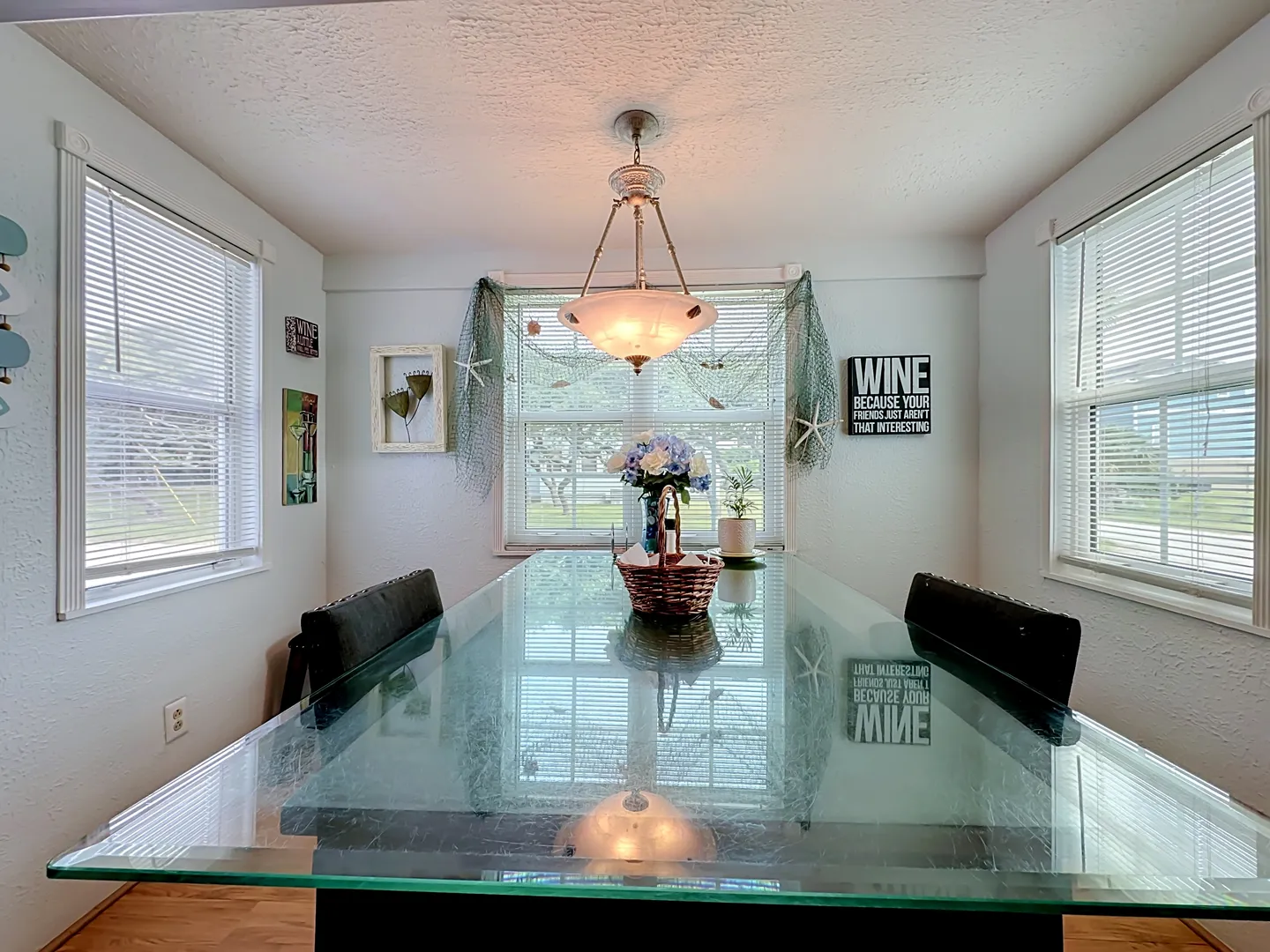Dining area with a long glass-top table, chairs, and coastal wall decorations framed by windows with blinds.