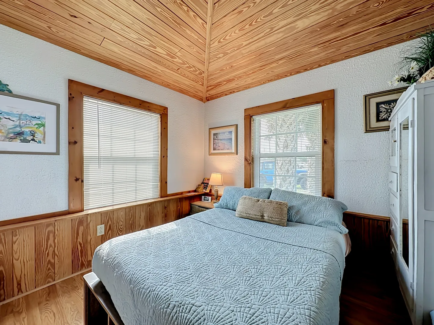 Bedroom with wood-paneled pyramid ceiling, wood wainscoting, light blue bed quilt, and two windows with blinds.