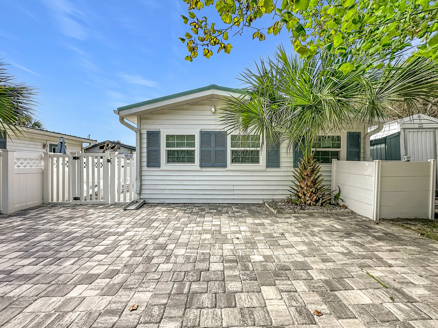 Exterior view of a white-sided single-story home with a paved stone patio, palm tree, and white vinyl fence.