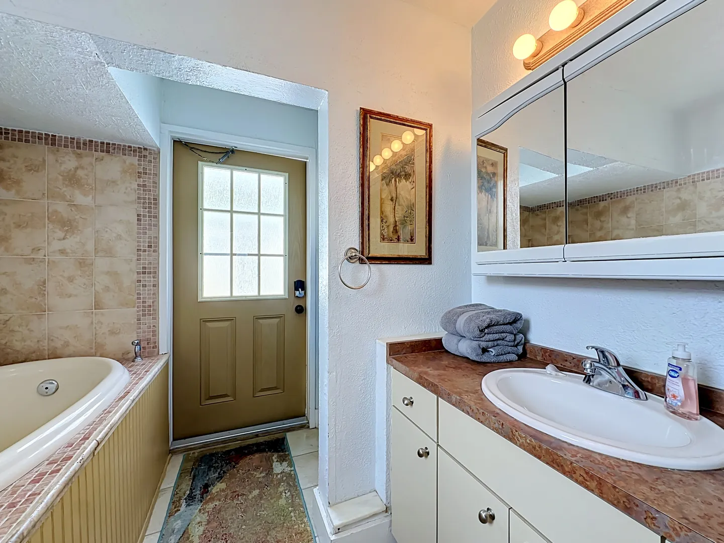 Bathroom showing a soaking tub tiled in tan ceramic on the left, a vanity with a sink on the right, and an exterior door with window panes at the back.