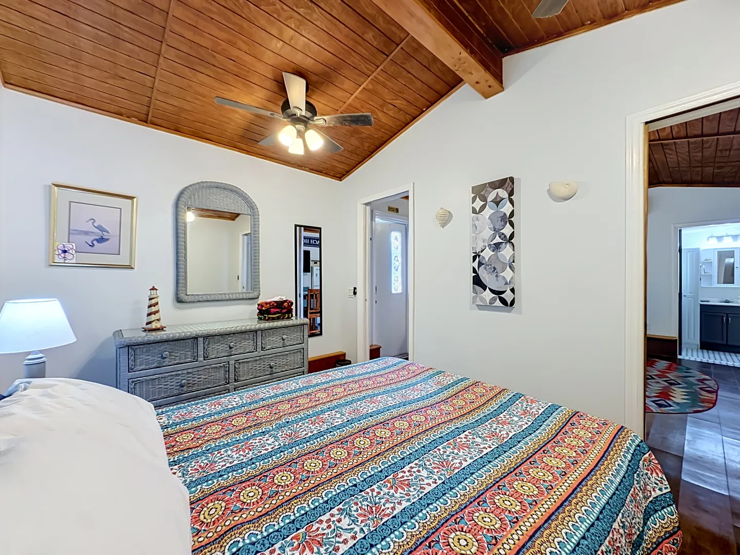 Bedroom with a sloped wood-plank ceiling, ceiling fan, colorful patterned bedspread, and woven dresser with a matching mirror.