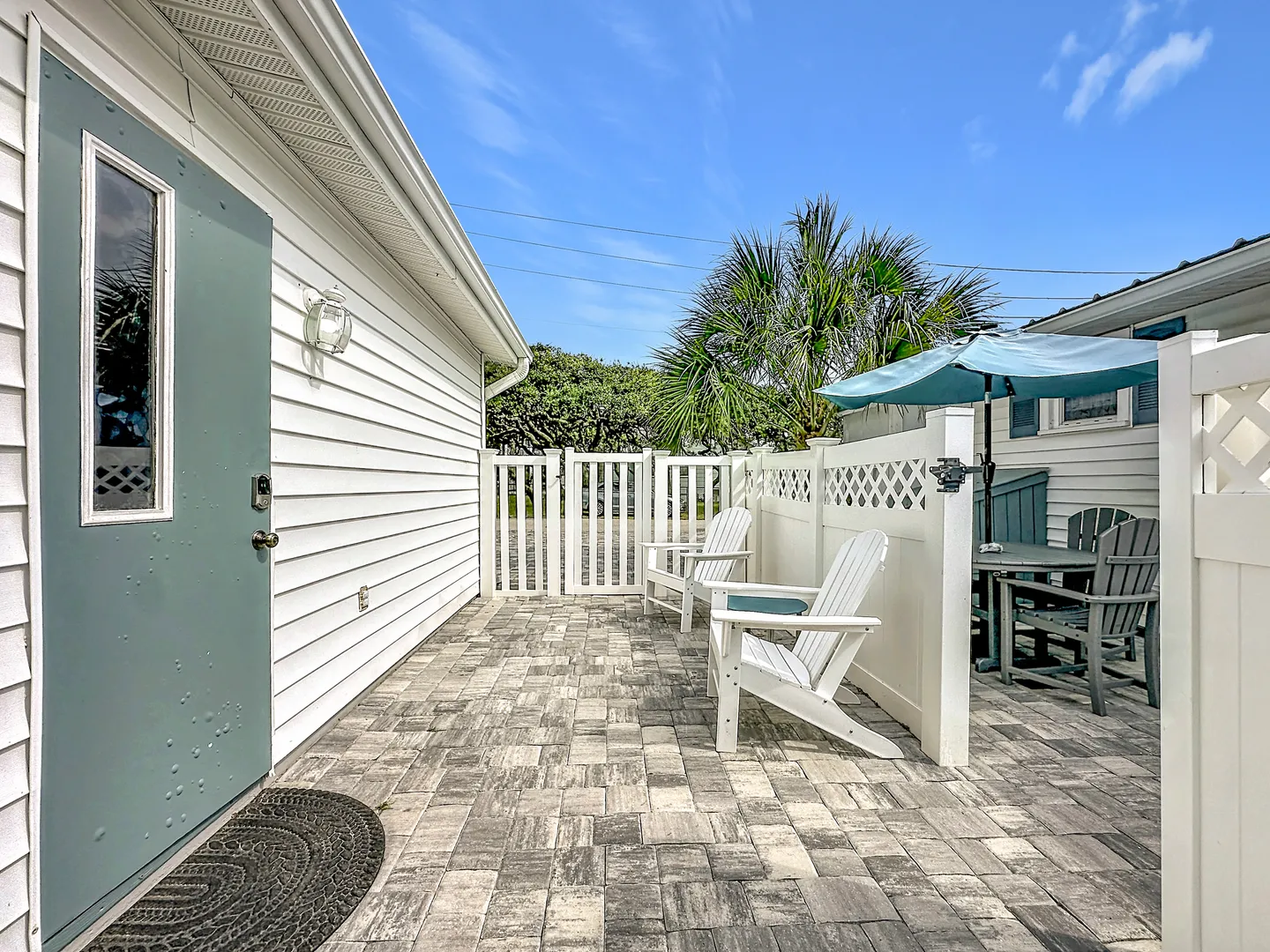 Paved outdoor patio area beside a house exterior with Adirondack chairs, an outdoor dining set under an umbrella, and white privacy fencing under a blue sky.