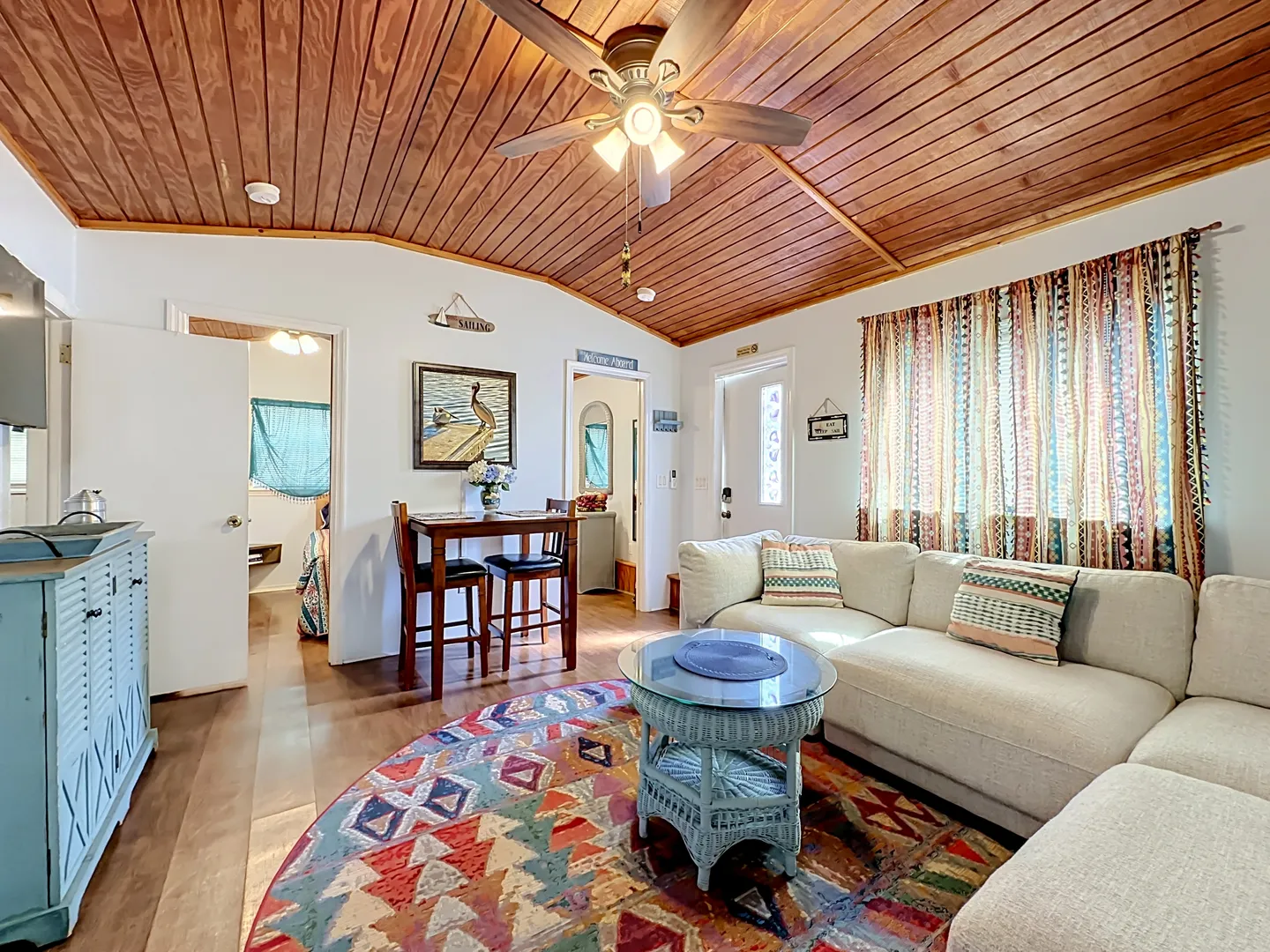 Living room with wood-paneled angled ceiling, ceiling fan, sectional sofa, round colorful rug, and a small dining table set.