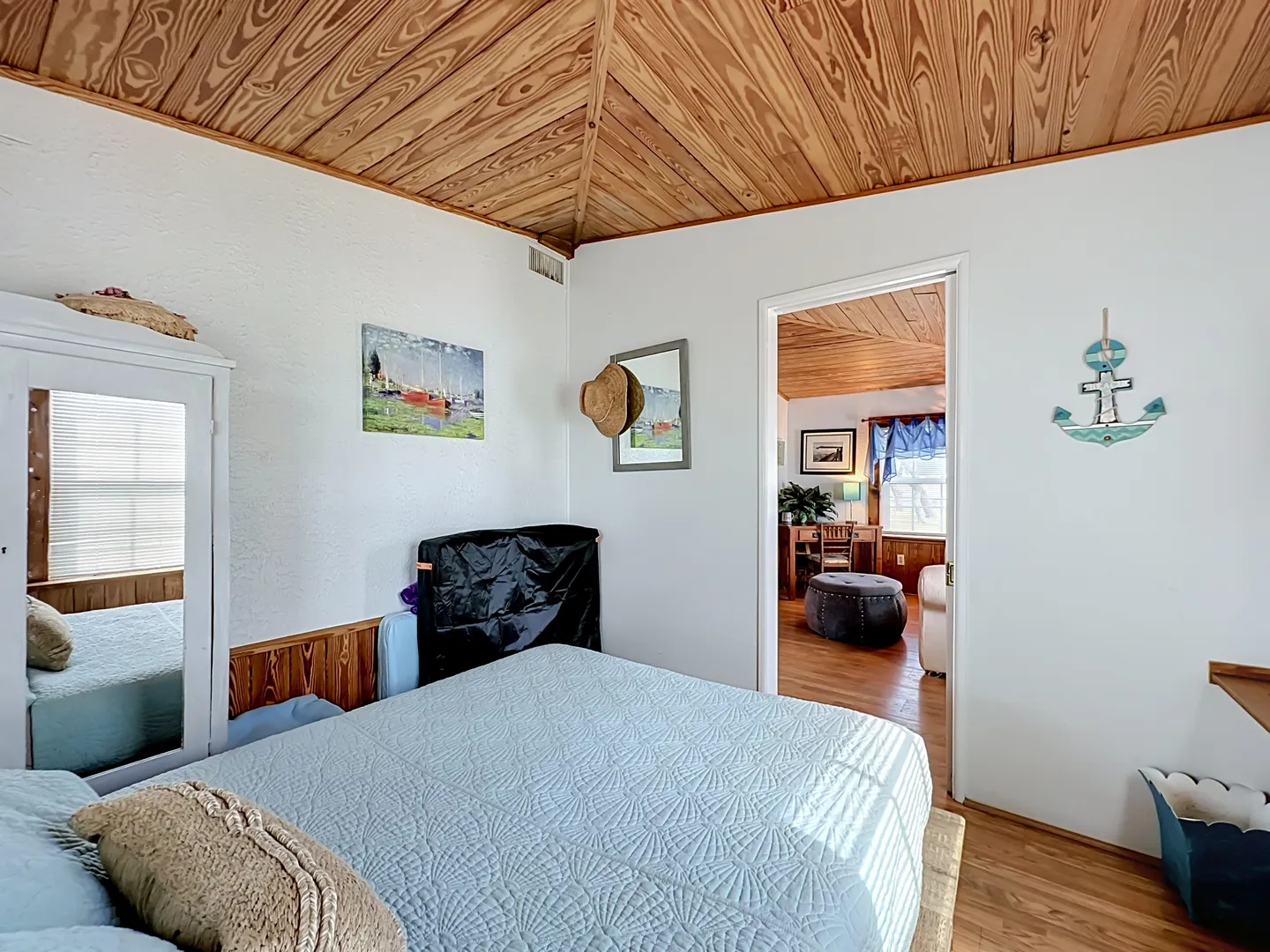 Bedroom with a vaulted wood-paneled ceiling, a made bed with a light blue quilt, white walls, and an open doorway looking into a living area.
