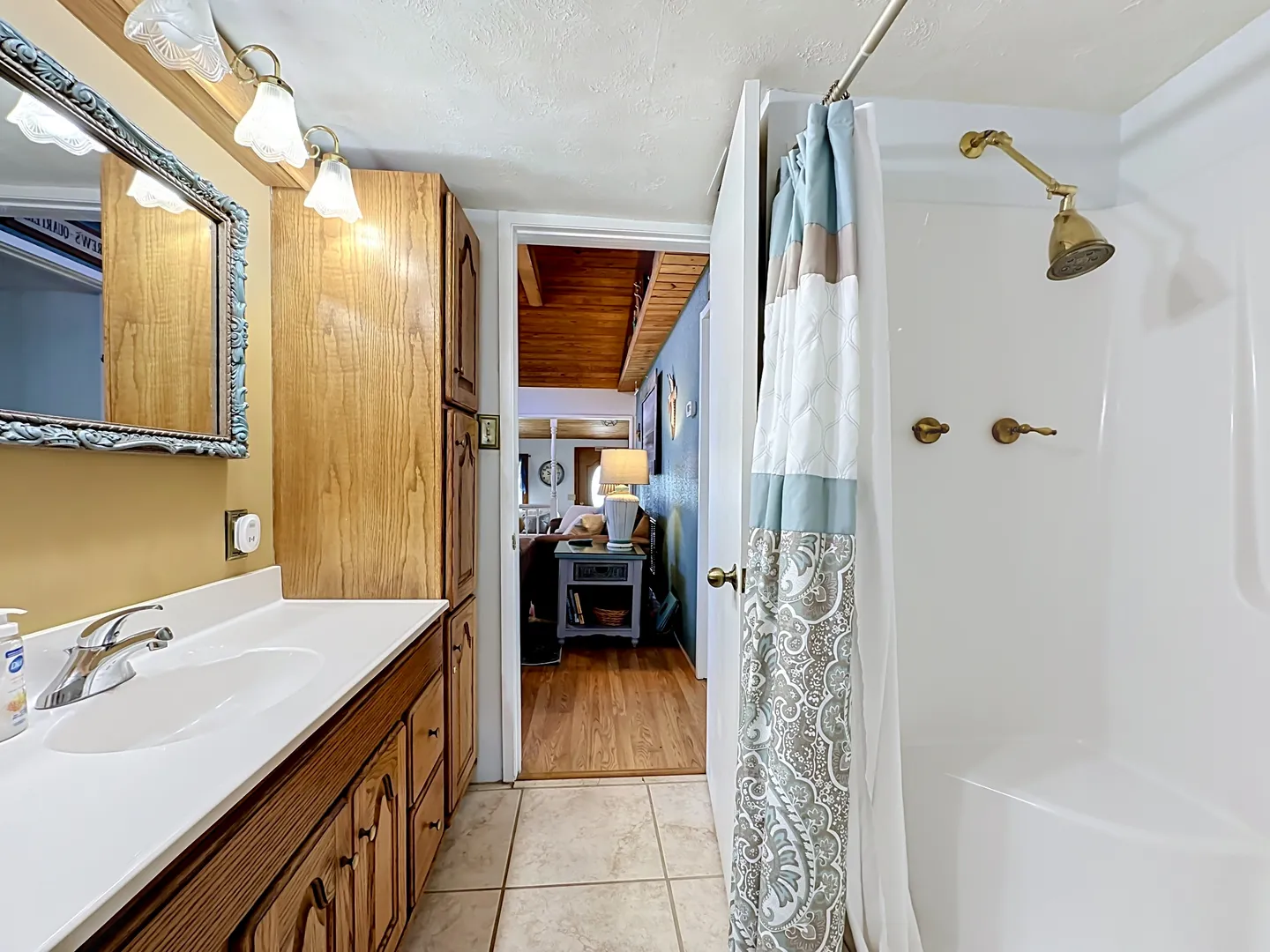 Bathroom showing a wooden vanity with a white countertop and mirror on the left and a white shower stall with a patterned curtain on the right.