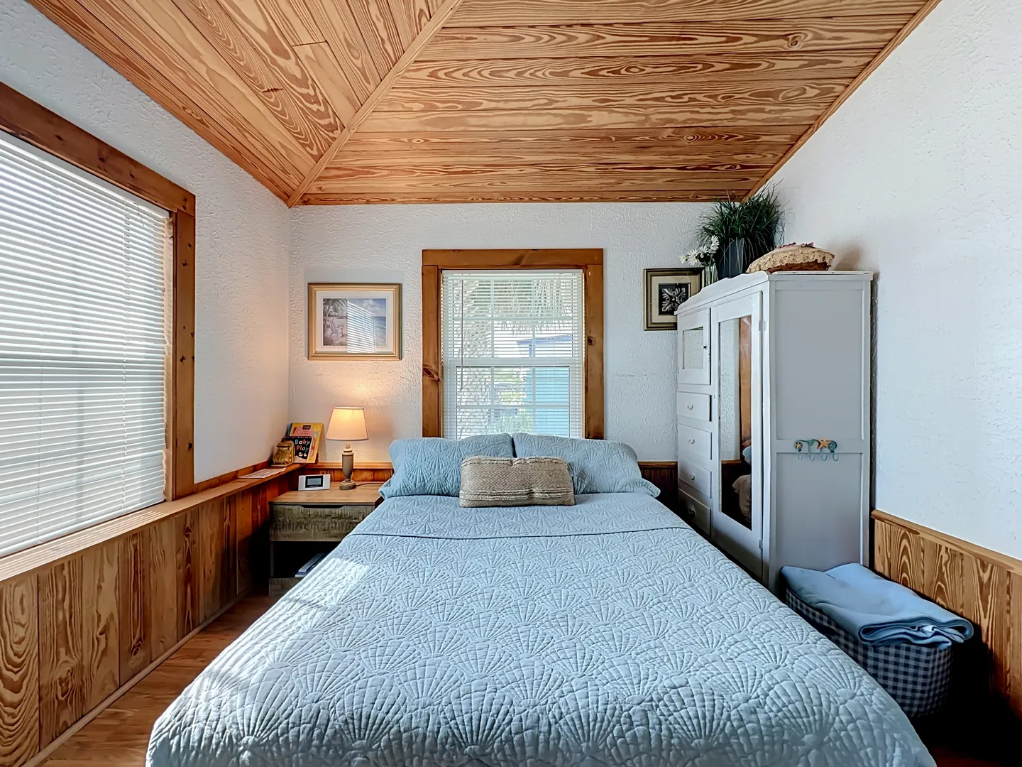 Bedroom with wood-paneled ceiling and wainscoting, featuring a made bed with a blue patterned quilt, a nightstand, and a white armoire.