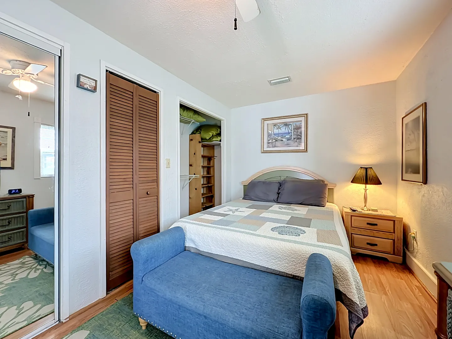 Bedroom with a bed made with a coastal quilt, a blue bench at the foot, wooden nightstand, and bifold closet doors.