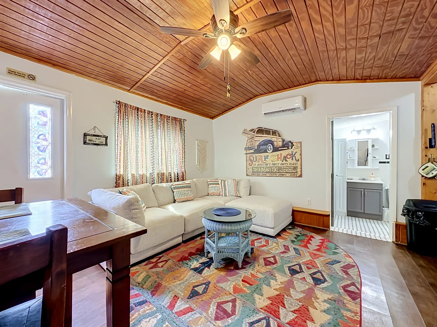 Living area featuring a sectional sofa, geometric patterned rug, light blue wicker coffee table, and wood-paneled ceiling with a fan.
