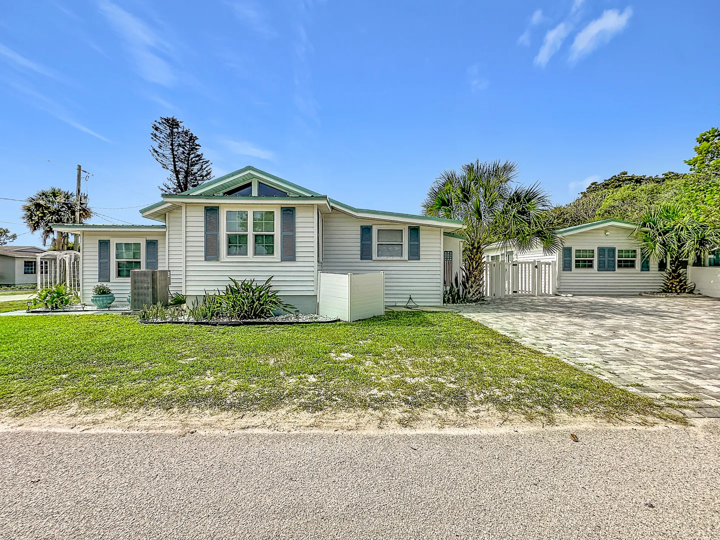 Exterior view of a single-story white siding coastal home with blue shutters, a paved driveway, and palm trees under a blue sky.