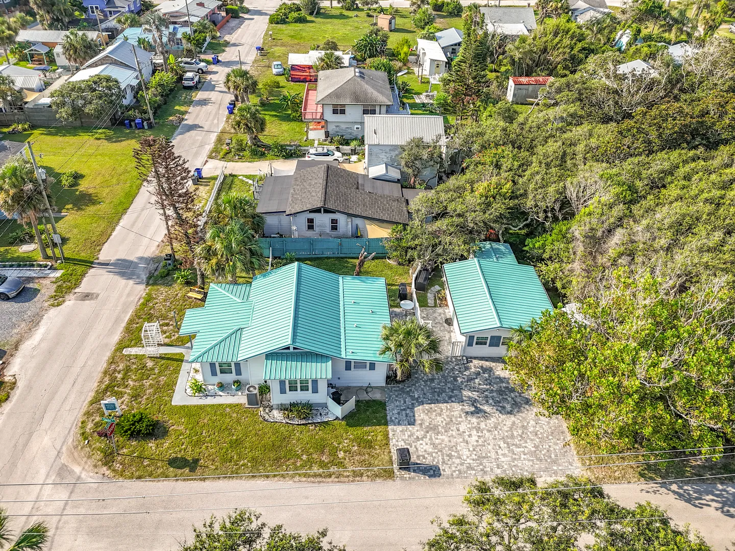 Aerial view of a coastal cottage property with turquoise metal roofs and a paved driveway surrounded by lush trees and neighboring homes.