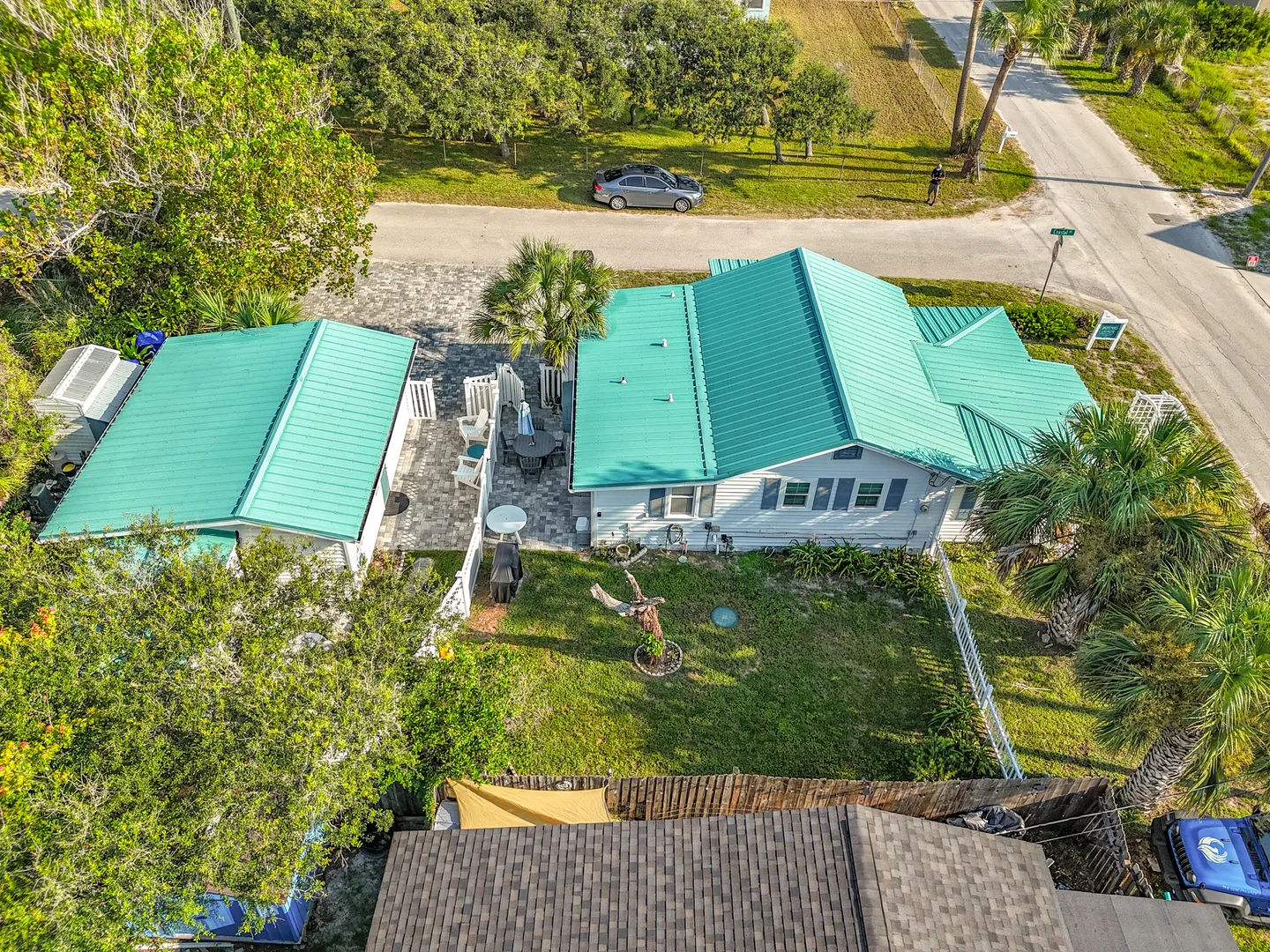Aerial view of a coastal property featuring light blue siding, a bright teal metal roof, a paved patio, and a grassy backyard.