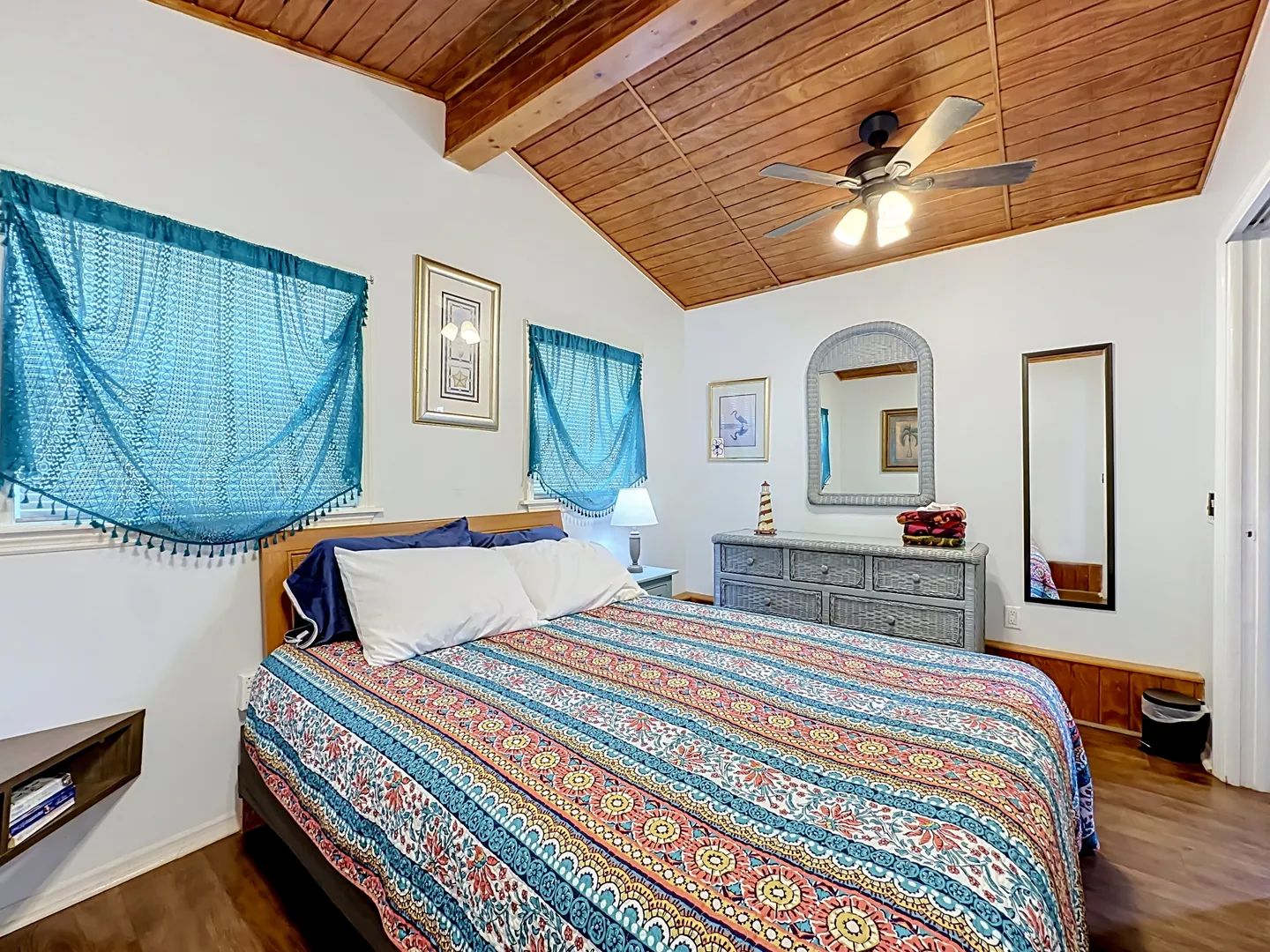 Bedroom with a patterned bedspread, wood plank ceiling with ceiling fan, teal window valances, and wicker dresser.