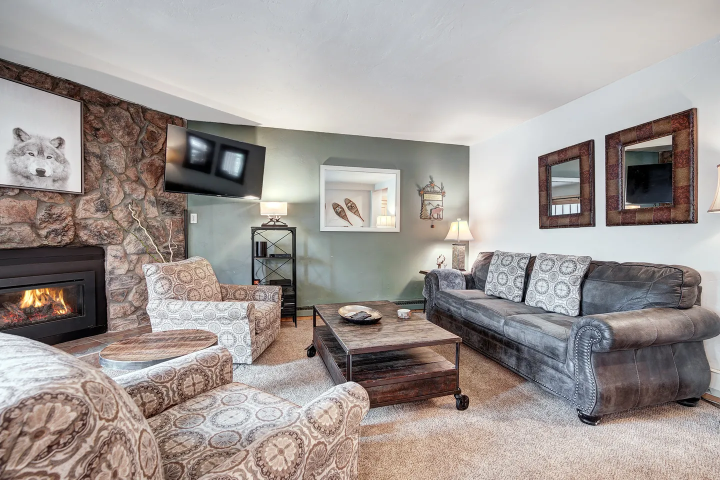 Living room with carpet, a stone fireplace with a fire, a wall-mounted TV, patterned armchairs, and a large sofa around a coffee table.