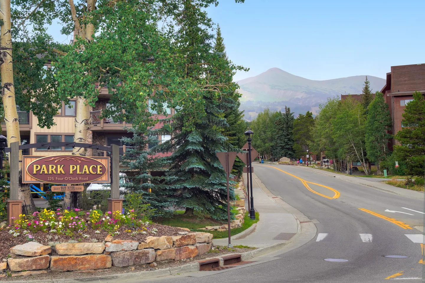 Street view showing the wooden entrance sign for Park Place at 325 Four O'Clock Road alongside landscaped flower beds, evergreen trees, and a mountain peak in the background.