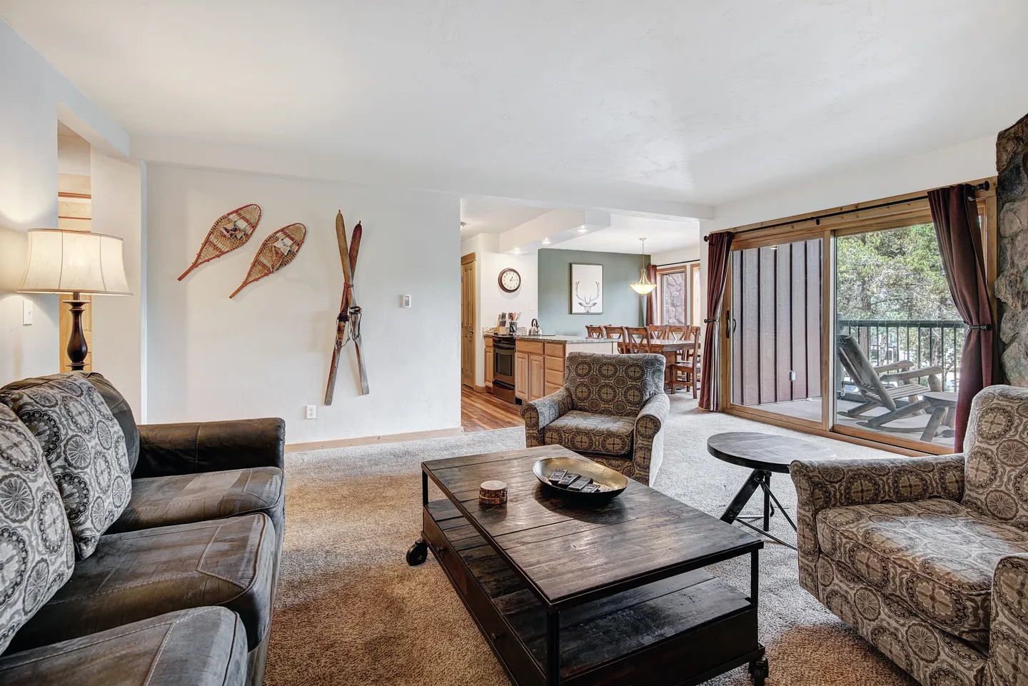 Living room with carpeted floor, rustic wall decor of skis and snowshoes, seating, coffee table, and sliding glass doors leading to a balcony.