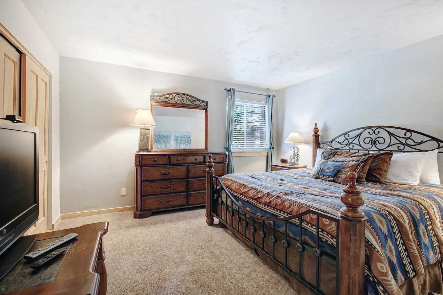 Bedroom featuring a wooden and wrought-iron bed frame, a matching wooden dresser with a mirror, a television on a stand, and carpeted flooring.