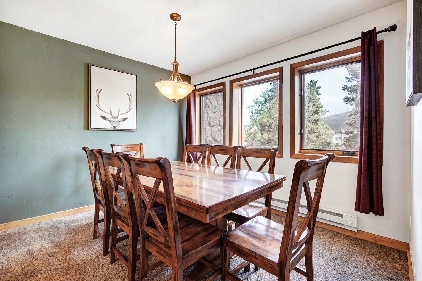 Dining area with a large wooden table and chairs, pendant lighting, carpeted floor, and windows showing trees outside.